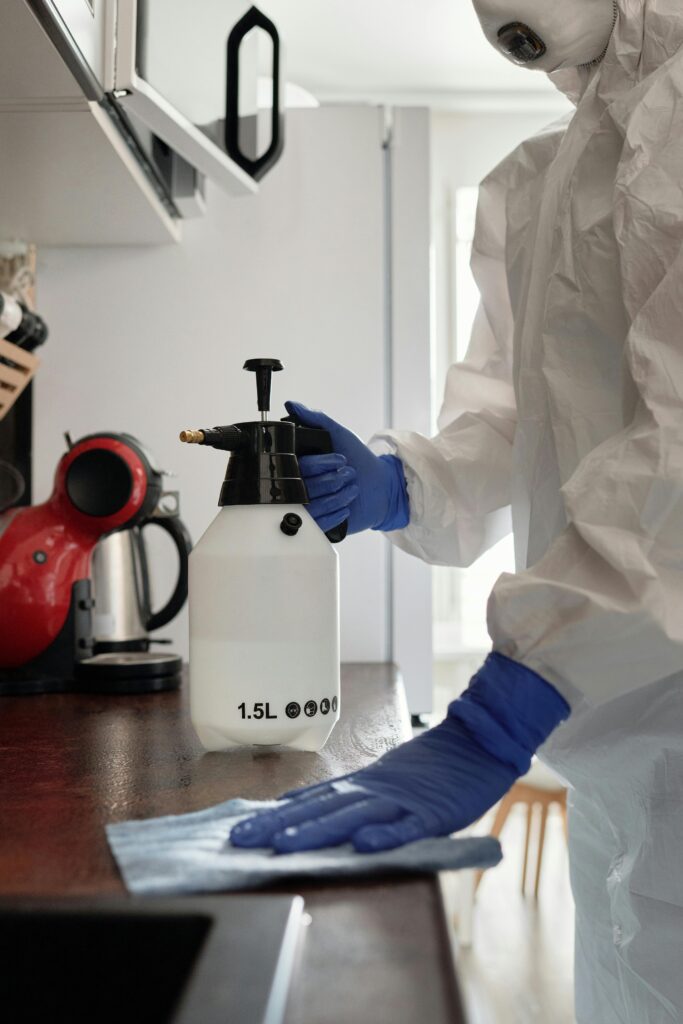 Person in protective suit cleaning a kitchen counter with spray bottle and gloves.